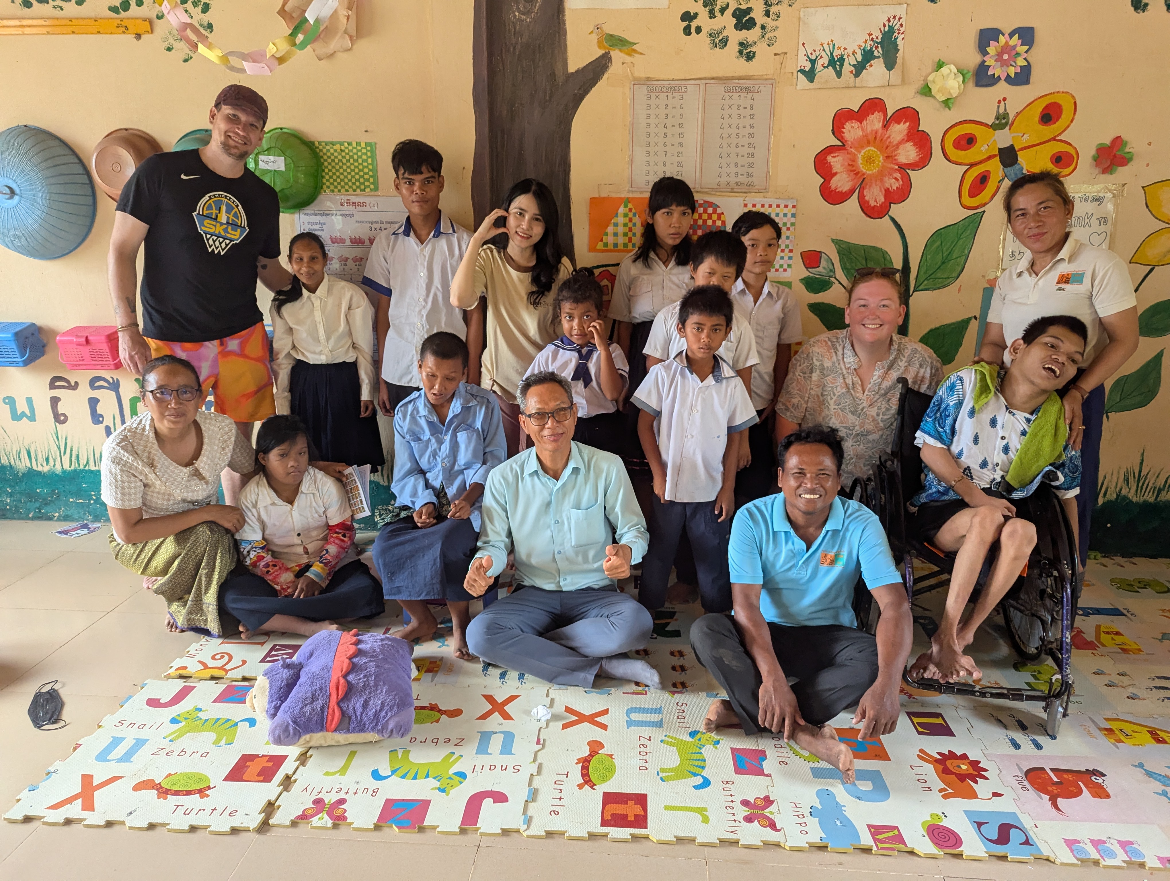 A group photo including Daniel, Hoa, Ashlyn, several teachers, and several students. The group is in a classroom filled with colorful murals, student art projects, and stands on a foam placemat patterned with letters of the alphabet and animals corresponding to each letter.