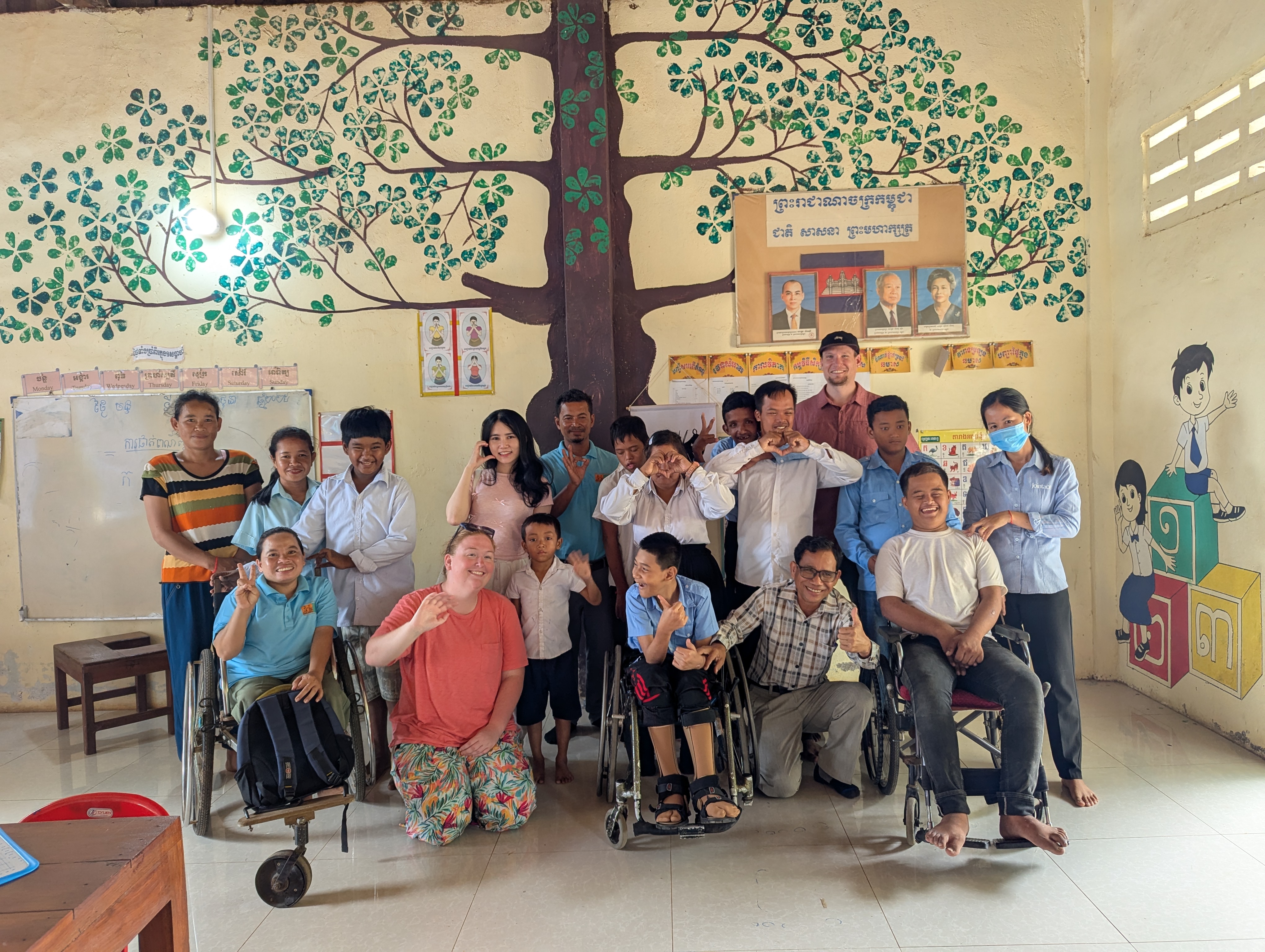 A group photo including Ashlyn, Hoa, Samnang, Daniel, several DDSP volunteers, and many students. The group is in a colorful classroom, and they are standing in front of a wall decorated with a large mural of a tree. Elsewhere in the classroom, there is a board showing pictures of Cambodian leaders, a mural of children sitting on oversized Khmer alphabet blocks, and educational signs.
