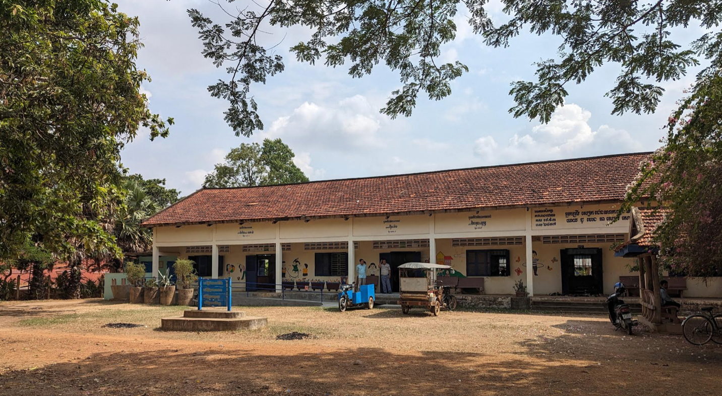 A single-story school building with a red-tiled roof and cream-colored walls in rural
Cambodia. The building has a row of open windows and doors, with educational mural and
Khmer script on the walls. An old tuk-tuk an small blue cart is parked in front. The yard is open
with dry grass, shaded by large trees, and is sunny.