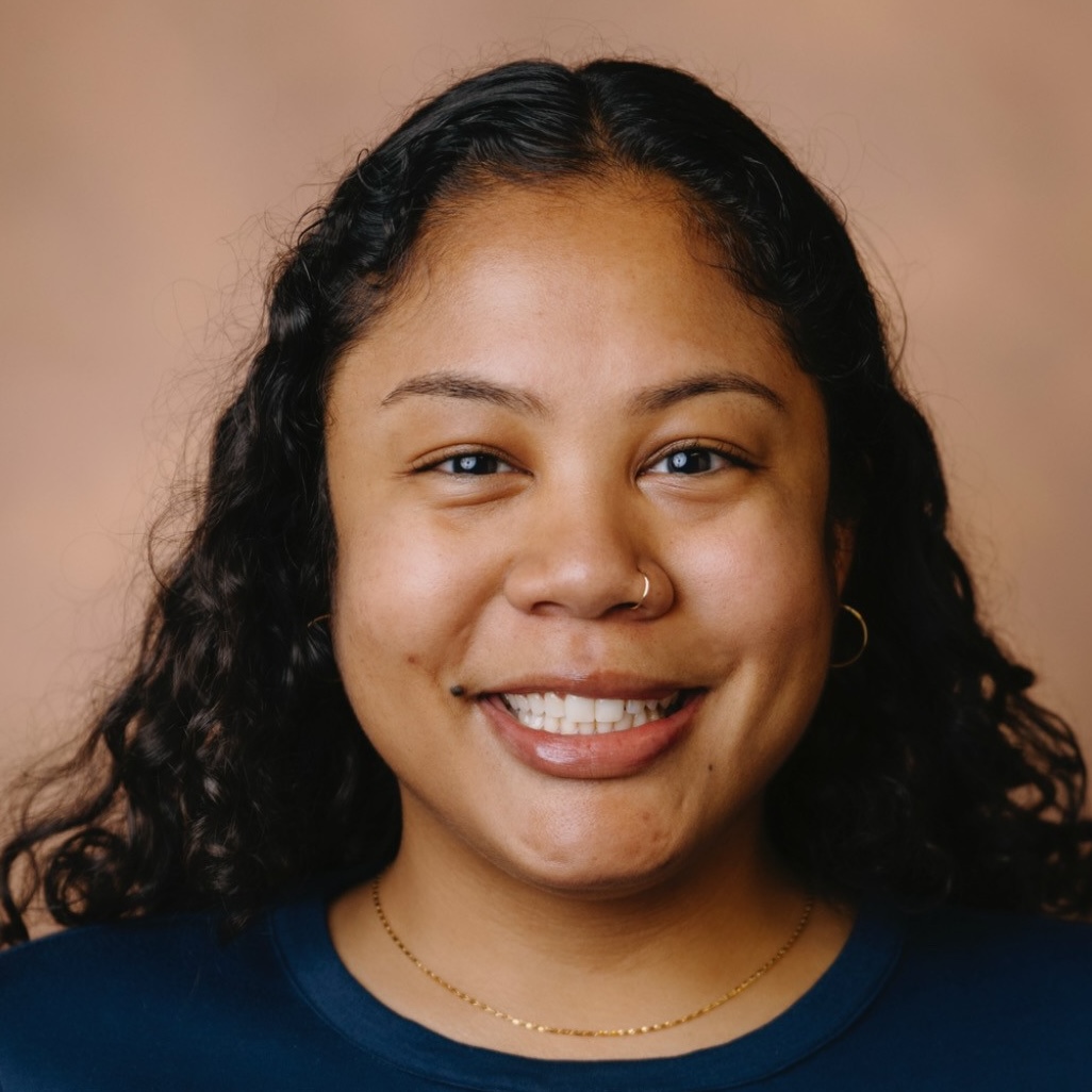 A headshot of Leanna. She has long curly dark hair, brown eyes, and is smiling. She is wearing a blue top.