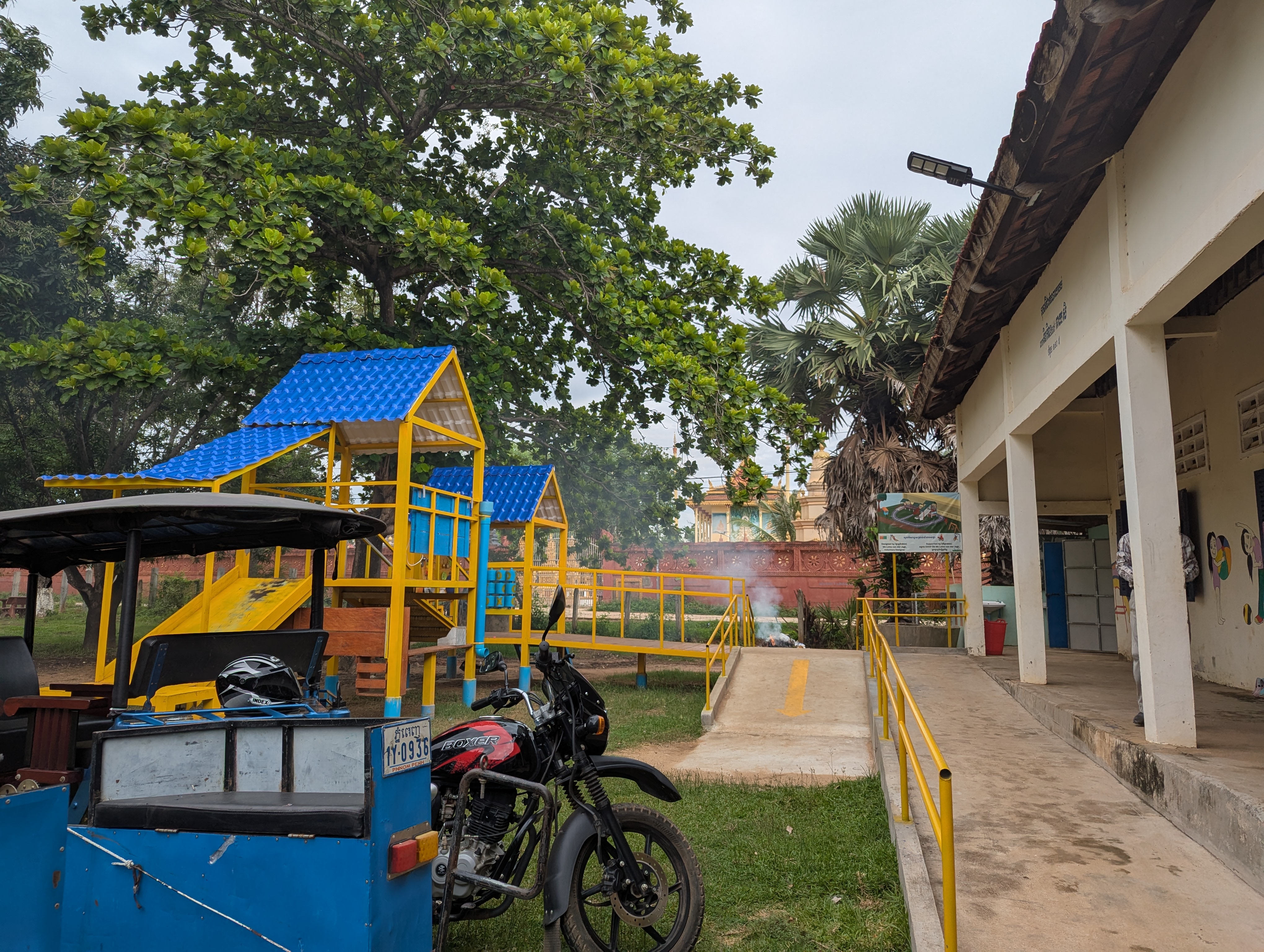 A school playground with a new bright blue and yellow playground, with a blue motorized tuk-tuk in the foreground.