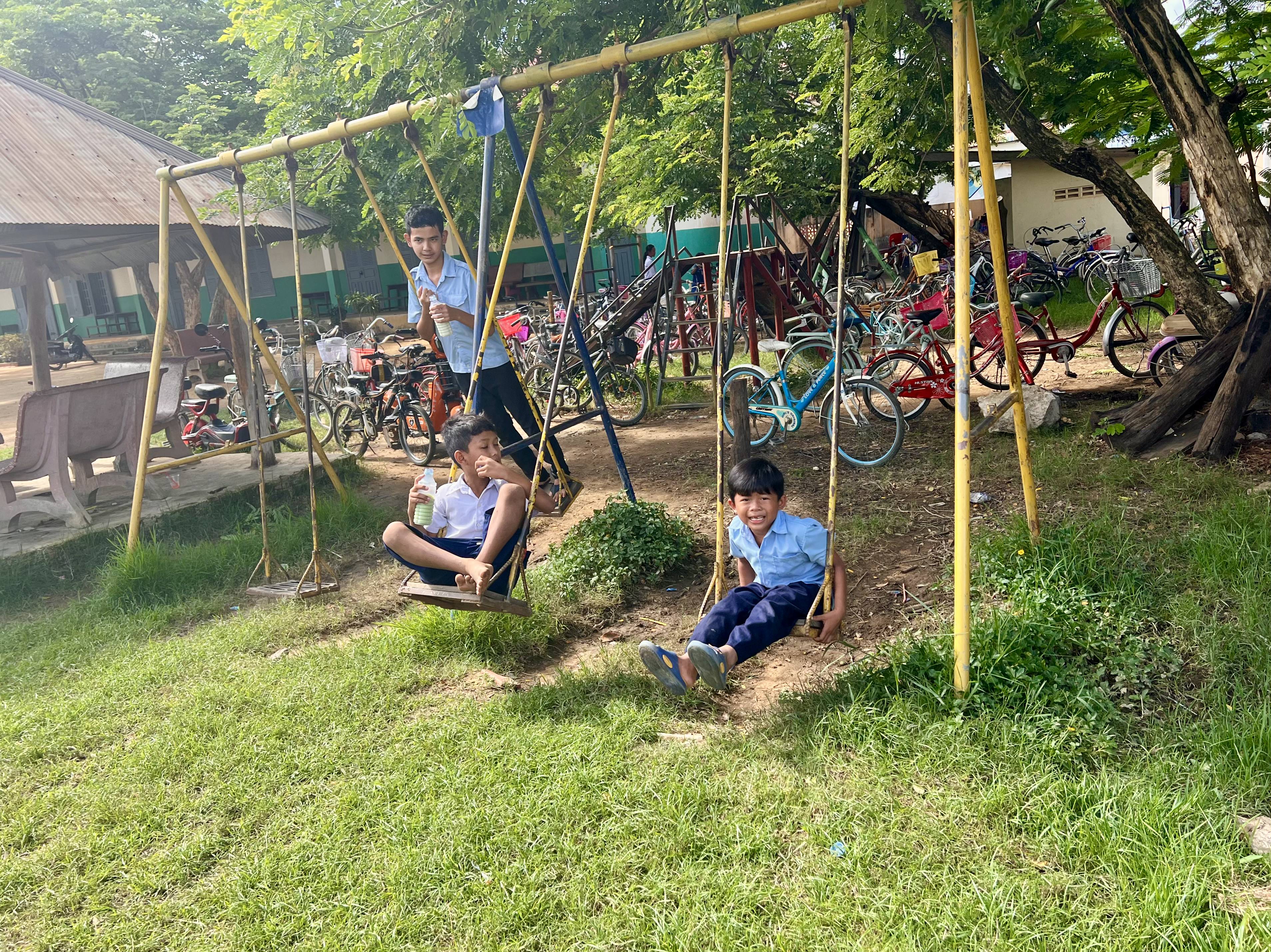 Three young students playing on a swingset outdoors on a sunny day. In the background, there are dozens of colorful new bikes.