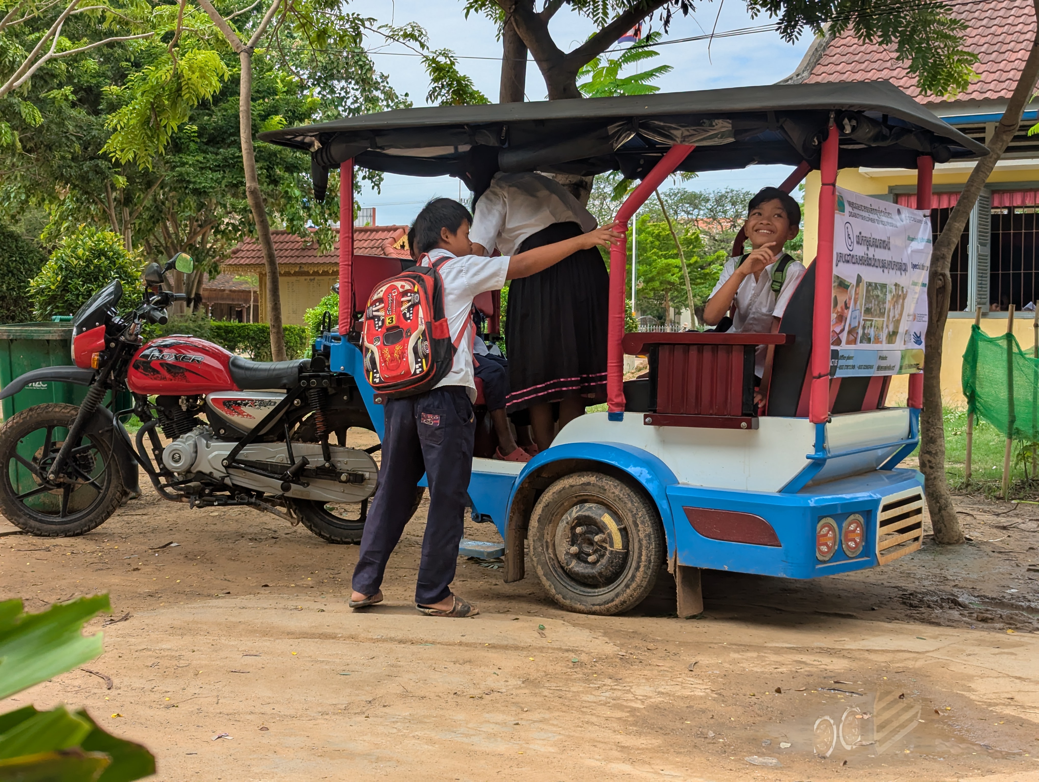 Four students board a new blue and red tuk-tuk in front of a school building.